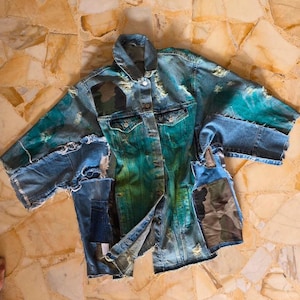 May include: A distressed denim jacket with a unique design. The jacket features a gradient of teal and blue hues, with patches of camouflage fabric. The sleeves and body have a ripped and frayed appearance, adding to its vintage aesthetic.