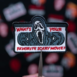 May include: Embroidered patch featuring the Ghostface mask from the movie Scream. The patch has the text "WHAT'S YOUR FAVORITE SCARY MOVIE?" in red and white. The patch is held by tweezers against a blurred background of similar patches.