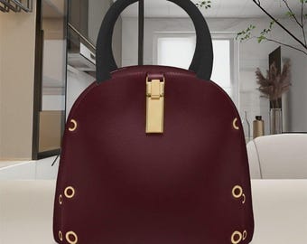 May include: A burgundy handbag with a black handle and gold-colored hardware. The bag has a rounded shape with gold eyelets along the sides and a gold clasp closure. The handle is a semi-circle shape.