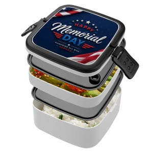 May include: A white, three-tiered bento box with a black lid featuring a Memorial Day design. The lid has a blue background with the text "Happy Memorial Day" and American flag imagery. The box is filled with food.