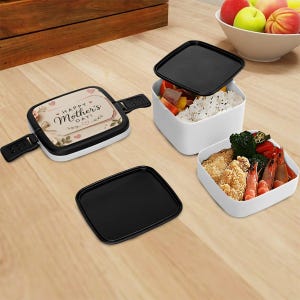 May include: A white and black lunch box with the text "Happy Mother's Day" and floral accents. Two additional white lunch containers with black lids are filled with food, including rice, vegetables, and fried items.