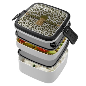 May include: A multi-tiered, white and black lunch box with a leopard print lid. The lid has a gold clasp and olive green accents. The lunch box is filled with rice and vegetables.