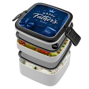 May include: A white and black Father's Day bento box with three compartments. The top lid features a blue graphic with the text "Happy Father's Day", a gift, a mustache, a crown, and a mug that says "Best Dad Ever". The bento box is filled with food.
