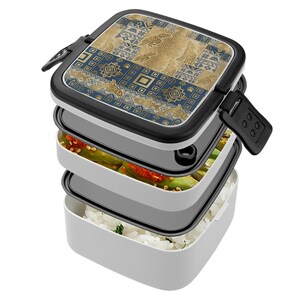 May include: A three-tiered, square lunch box with a black lid featuring a geometric pattern in gold and navy blue. The box is open, revealing layers of food: rice, vegetables, and other ingredients. The box has a black clasp.