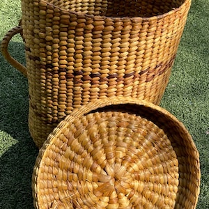 May include: A large, woven basket with a lid, crafted from natural fibers. The basket is cylindrical with a handle and features a lid. The basket is approximately 24 inches tall and 18 inches in diameter, with a lid that is 18 inches in diameter.