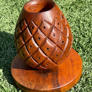 May include: A handcrafted wooden pineapple-shaped container with a diamond pattern and small holes. The container sits on a round wooden base. The wood has a warm, reddish-brown color.