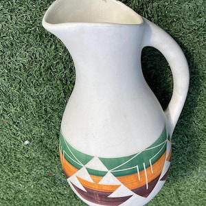 May include: A white ceramic pitcher with a curved handle and a wide spout. The base features a geometric pattern in green, orange, brown, and white. The pitcher is likely used for serving beverages.