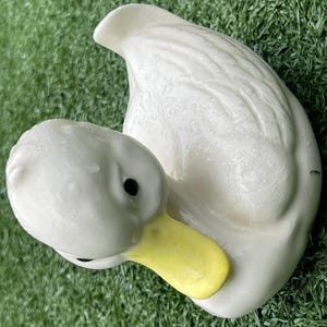 May include: A white ceramic duck figurine with a yellow beak and black eyes. The duck is in a resting position, with its head turned towards its body. The figurine has a glossy finish and is set against a green background.