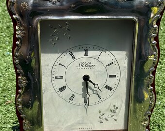 May include: A rectangular clock with a silver-colored frame and a dark red border. The clock face is white with black Roman numerals and hands. The clock face has the text "R. Carr Clockmaker England".