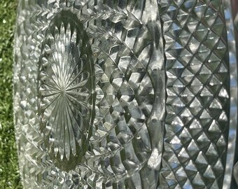 May include: A clear, round glass serving platter with a detailed diamond and starburst pattern. The edges are slightly raised, and the surface reflects light, creating a sparkling effect. Ideal for serving appetizers or desserts.