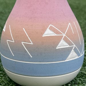 May include: A decorative vase with a gradient of pink to orange to blue. White geometric designs are etched into the vase. The base is a light green color. The vase is sitting on green grass.