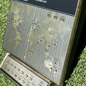May include: Vintage Sunbeam World Time Alarm Clock with a map design. Features a digital display, time zone indicators, and control buttons. The clock has a silver and black color scheme.