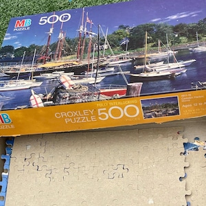 May include: A partially assembled 500-piece jigsaw puzzle featuring a harbor scene with sailboats and a schooner. The puzzle box, labeled "Croxley Puzzle," shows the completed image. The puzzle is for ages 12 and up.