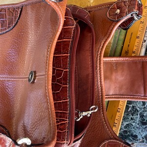 May include: A brown leather handbag with a textured, crocodile-like pattern. The bag features a zippered compartment, a silver-colored clasp, and decorative stitching. The bag has a handle and a shoulder strap.
