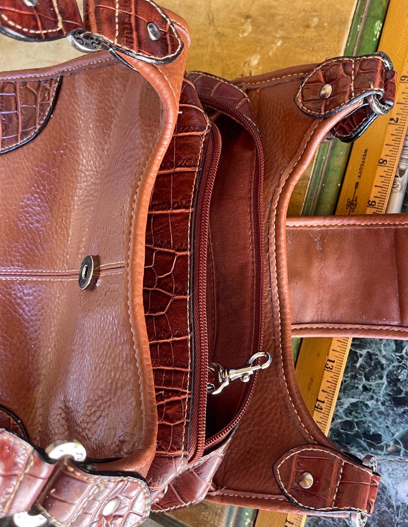 May include: A brown leather handbag with a textured, crocodile-like pattern. The bag features a zippered compartment, a silver-colored clasp, and decorative stitching. The bag has a handle and a shoulder strap.