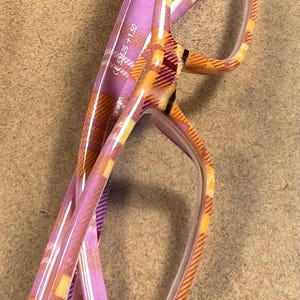 May include: Reading glasses with a plaid pattern in shades of purple, orange, and yellow. The frames have a rectangular shape and the lenses are clear. The glasses are marked with "CE 53626 +1.50".
