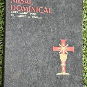 May include: A dark gray book titled "MI MISAL DOMINICAL" with red text. The cover features a cross and chalice design. The book includes text in Spanish, and is on a green surface.