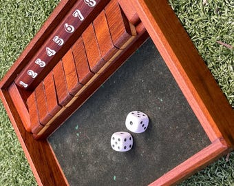 May include: A wooden shut-the-box game with a dark green felt playing surface. The game features wooden tiles numbered 1-9, and two white dice with black dots. The frame is made of stained wood.