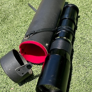 May include: A black camera lens with a 400mm focal length, alongside its black carrying case and lens hood. The case is cylindrical with a red interior. The lens is resting on green grass.