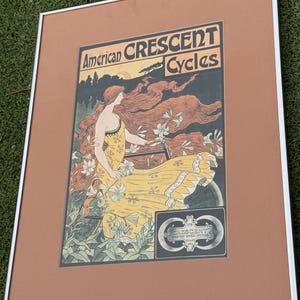 May include: A framed vintage-style poster titled American Crescent Cycles featuring an illustration of a person with long flowing red hair wearing a yellow dress while riding a bicycle amidst white lilies, with a black and white logo in the corner.