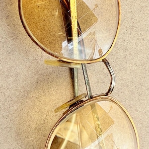 May include: Vintage round eyeglasses with gold-tone frames and yellow-tinted lenses. The glasses have a double bridge and a small, rectangular detail at the top of the frame. The earpieces are white and the lenses have some cracks.
