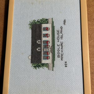 May include: Needlepoint of the Biddle House on Mackinac Island, 1986. The house has a black roof, white walls, and red window accents. The needlepoint is set in a light blue background and framed in a beige border.