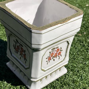 May include: A square, white ceramic planter with a green trim and floral design. The planter has a pedestal base and a green fabric lining the top rim. The floral design features orange and yellow flowers.