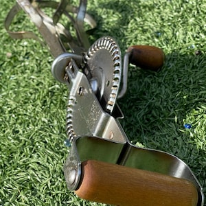 May include: Vintage hand-crank egg beater with a wooden handle and a gear-driven mechanism. The beater is made of metal and features a series of looped wires for whisking. The wooden handle is a warm brown color. The beater is resting on a bed of green grass.