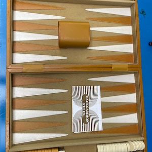 May include: Open backgammon set in a wooden case with tan and white playing surface. Includes a tan leather dice cup, dice, and game pieces. A small booklet with the text "BACKGAMMON RULES" is also included.