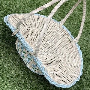 May include: A white wicker basket with a light blue trim and handle. The basket has a floral design on the side and is sitting on a green surface. The basket has a woven design and a sturdy handle.