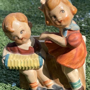 May include: A vintage ceramic planter featuring two figures: a boy playing an accordion and a girl. The boy wears a maroon vest and orange socks. The girl is in an orange dress and blue scarf. The planter has a cream-colored base.
