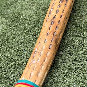 May include: A cylindrical, light brown wooden object with numerous small, dark holes. The bottom end is wrapped with a colorful band of turquoise, red, and yellow stripes. The object is lying on a green surface, with a measuring tape visible.