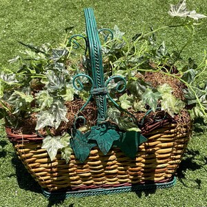 May include: A decorative wicker basket with a green metal handle and accents. The basket is filled with faux ivy and moss. The handle features decorative swirls and leaf-shaped accents. The basket is sitting on a green surface.