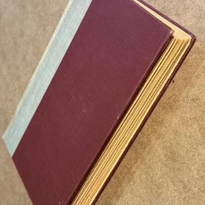 May include: A hardcover book with a burgundy cover and a light blue spine. The pages are yellowed with age, and the book is slightly angled, revealing the side of the pages. The book is likely vintage or antique.