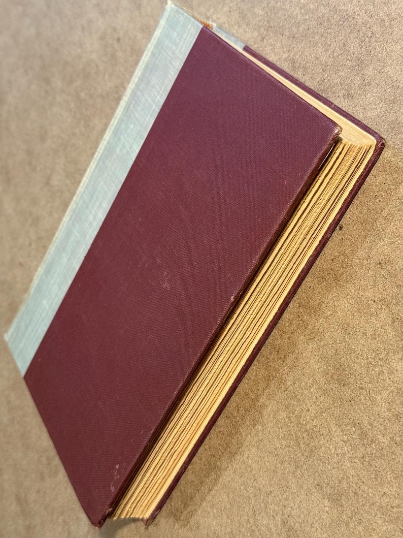 May include: A hardcover book with a burgundy cover and a light blue spine. The pages are yellowed with age, and the book is slightly angled, revealing the side of the pages. The book is likely vintage or antique.
