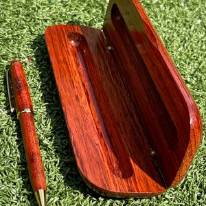 May include: A handcrafted wooden pen and pen case. The pen is a rich reddish-brown with gold-colored accents. The open case, also reddish-brown, has a curved design with a groove to hold the pen. The pen and case are on a green surface.