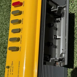 May include: A bright yellow stereo radio cassette recorder featuring a black cassette deck door, a red record button, and several grey control buttons labeled play, rewind, f fwd, stop/eject, and pause. The device includes a radio tuner dial.