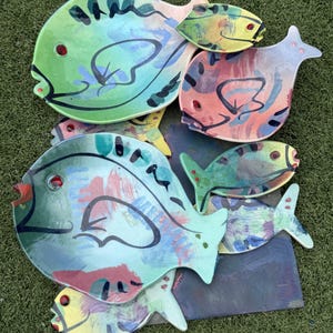 May include: A collection of seven ceramic fish-shaped plates and decorative pieces featuring hand-painted abstract designs in vibrant shades of green, blue, red, and yellow, with bold black line work and small red circular accents for eyes.