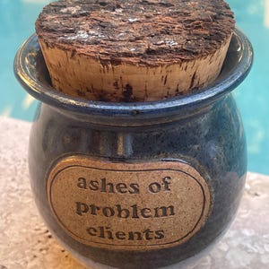 May include: A small, blue ceramic jar with a cork lid. The jar has a brown label with the words "ashes of problem clients" in a serif font. The jar is sitting on a stone surface.
