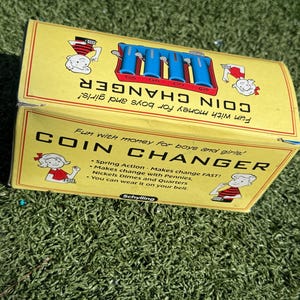 May include: A vintage yellow coin changer box with blue coin tubes and the words "COIN CHANGER" in large black letters. The box has illustrations of children and text that reads "Fun with money for boys and girls!"