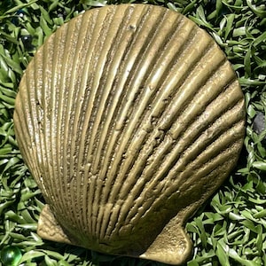 May include: A metallic, gold-toned scallop shell object with a textured, ribbed surface. The shell features a fan-shaped design with distinct, raised vertical ridges radiating from the base to the rounded outer edge, showing a weathered finish.