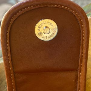 May include: Close-up of a brown leather handbag flap with a magnetic closure. The closure features a circular metal piece with the numbers "AND06631889" and "NO.6633918" engraved on it. The stitching is visible around the edges.