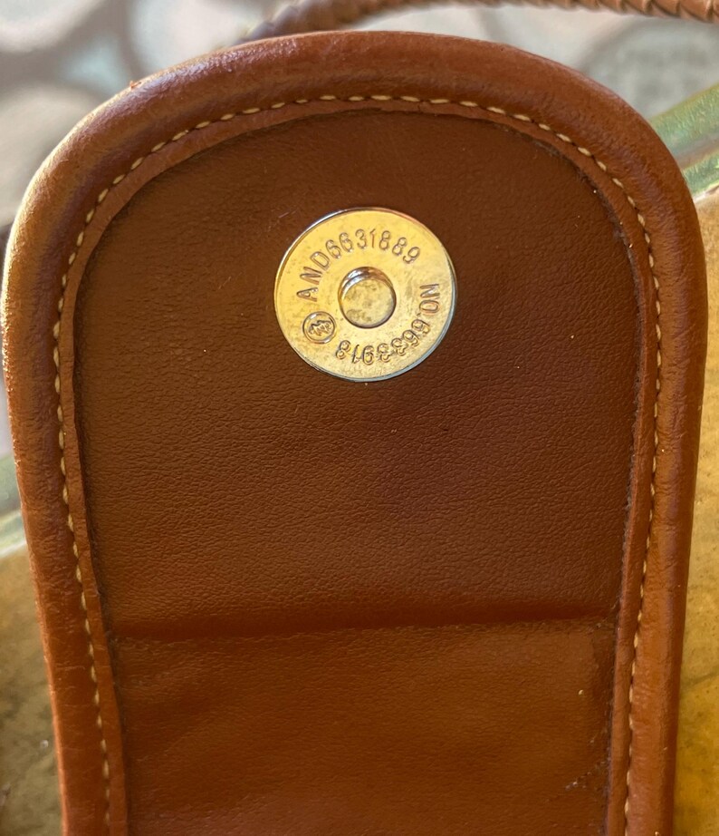 May include: Close-up of a brown leather handbag flap with a magnetic closure. The closure features a circular metal piece with the numbers "AND06631889" and "NO.6633918" engraved on it. The stitching is visible around the edges.