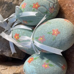 May include: Four light blue decorative Easter eggs with red and yellow floral designs and white ribbon bows. The eggs have a textured surface and are arranged on a stone background.