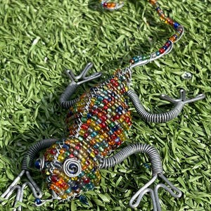 May include: A handcrafted lizard sculpture made of colorful beads and silver wire. The lizard's body is covered in a mosaic of red, yellow, green, and blue beads. The legs and tail are made of coiled wire. The sculpture is displayed on a bed of green grass.