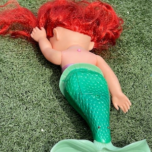 Pode incluir: Boneca de sereia com cabelo ruivo, tronco rosa e cauda verde. A boneca est&aacute; deitada sobre uma superf&iacute;cie verde. A boneca sereia tem uma barbatana caudal verde claro.