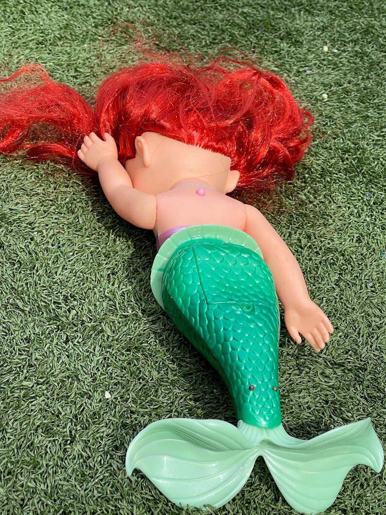 Pode incluir: Boneca de sereia com cabelo ruivo, tronco rosa e cauda verde. A boneca est&aacute; deitada sobre uma superf&iacute;cie verde. A boneca sereia tem uma barbatana caudal verde claro.
