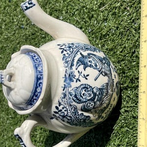 May include: White ceramic teapot with blue floral and figural designs. The teapot has a curved spout, a handle, and a lid. The blue designs include figures and floral patterns. The teapot is on a green surface.