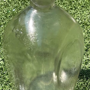 May include: A clear, antique glass bottle with a rounded body and a narrow neck. The bottle has a slightly greenish tint and appears to be made of thick glass. The bottle is sitting on a green surface.