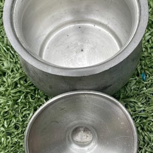 May include: A round, open, aluminum pot with a matching lid. The pot has a wide rim and a slightly textured surface. The lid is circular with a central knob. The pot and lid are sitting on a bed of green grass.
